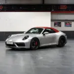 Porsche 992 911 Carrera GTS Cabriolet von 2023 in Silber mit geschlossenem rotem Verdeck, seitliche Frontansicht in einer Ausstellungshalle.