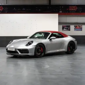 Porsche 992 911 Carrera GTS Cabriolet von 2023 in Silber mit geschlossenem rotem Verdeck, seitliche Frontansicht in einer Ausstellungshalle.