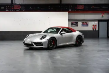 Porsche 992 911 Carrera GTS Cabriolet von 2023 in Silber mit geschlossenem rotem Verdeck, seitliche Frontansicht in einer Ausstellungshalle.