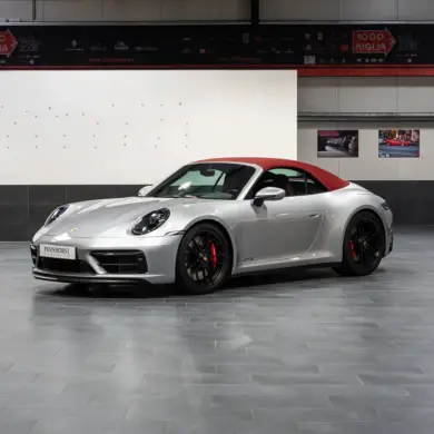 Porsche 992 911 Carrera GTS Cabriolet von 2023 in Silber mit geschlossenem rotem Verdeck, seitliche Frontansicht in einer Ausstellungshalle.