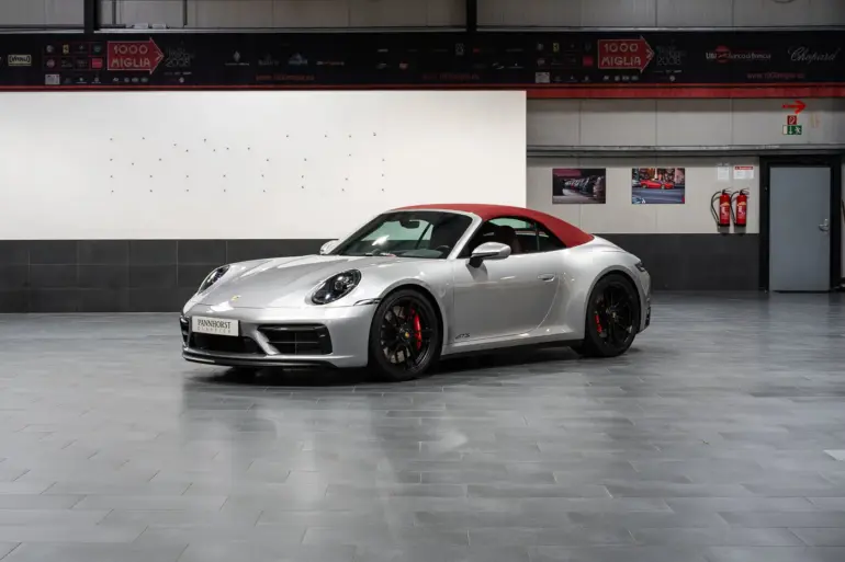 Porsche 992 911 Carrera GTS Cabriolet von 2023 in Silber mit geschlossenem rotem Verdeck, seitliche Frontansicht in einer Ausstellungshalle.