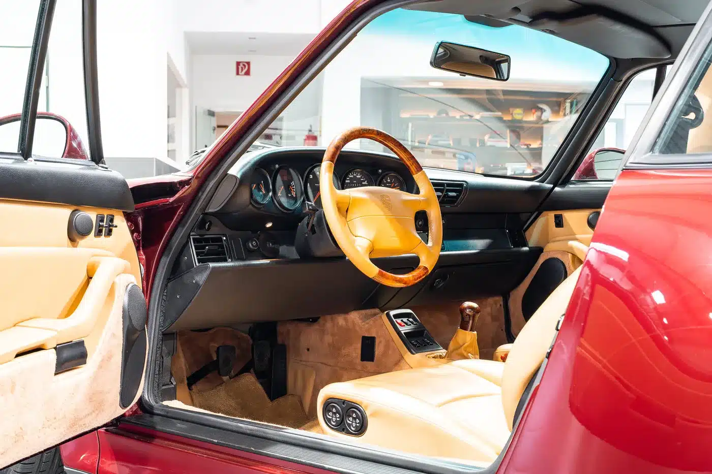 Cockpit des Porsche 993 Carrera 4S mit beigem Leder, Holzlenkrad und geöffneter Tür in Arena Rot Metallic