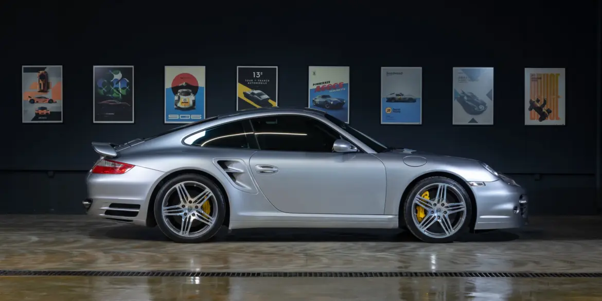 Silberner Porsche 997 Turbo 2006 seitlich in einer Halle, gut beleuchtet, gelbe Bremssättel sichtbar, Autos als Poster im Hintergrund.