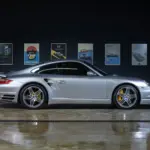 Silberner Porsche 997 Turbo 2006 seitlich in einer Halle, gut beleuchtet, gelbe Bremssättel sichtbar, Autos als Poster im Hintergrund.