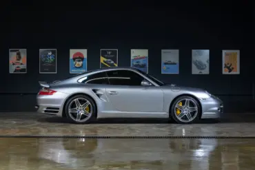Silberner Porsche 997 Turbo 2006 seitlich in einer Halle, gut beleuchtet, gelbe Bremssättel sichtbar, Autos als Poster im Hintergrund.