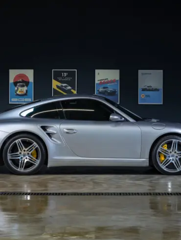 Silberner Porsche 997 Turbo 2006 seitlich in einer Halle, gut beleuchtet, gelbe Bremssättel sichtbar, Autos als Poster im Hintergrund.