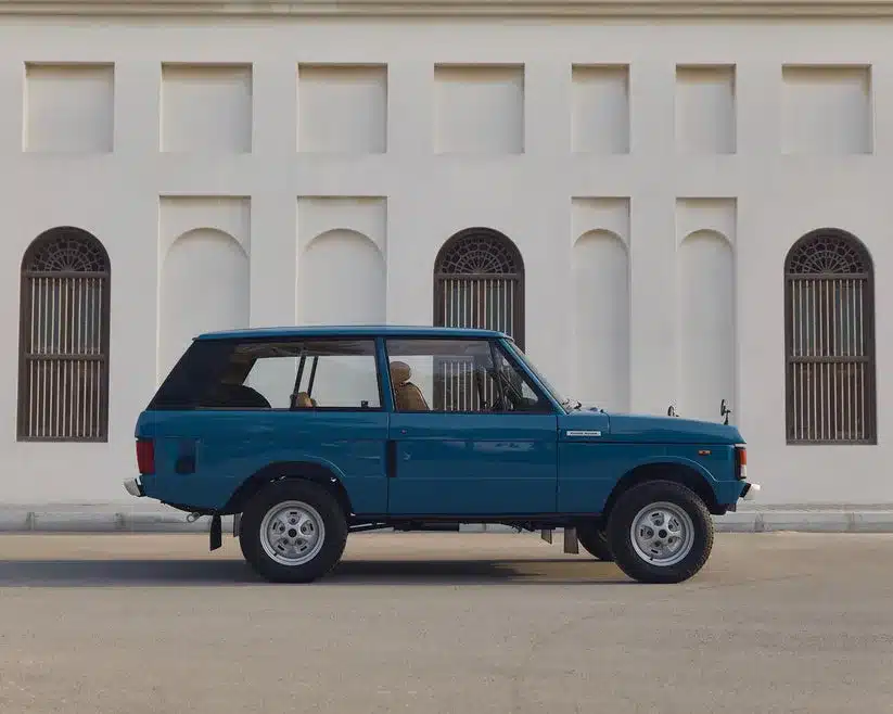 Restaurierter Range Rover Classic von 1975 in Tuscan Blue, Seitenansicht vor heller Wand mit Bogengittern.