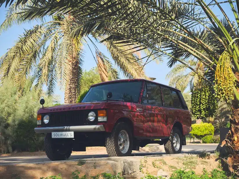 Range Rover Classic von 1976 in rotem Lack (Masai Red) im Freien unter Palmen, Ansicht schräg von vorne.