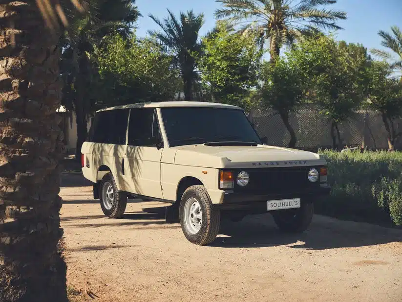 Range Rover Classic 1981 in Sahara Dust Lackierung auf sandigem Weg mit Palmen im Hintergrund.