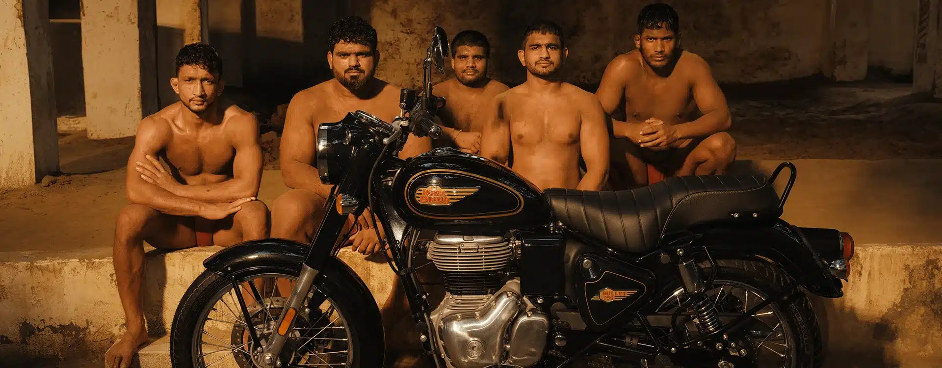 Die Royal Enfield Bullet 350, seitlich aufgenommen, mit fünf muskulösen Ringern im Hintergrund.