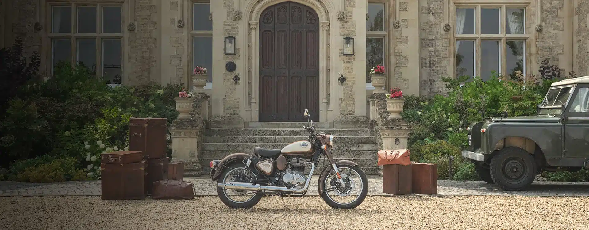 Royal Enfield Classic 350 Motorrad in Medallion Bronze, geparkt vor einem Herrenhaus, daneben Reisegepäck und ein Oldtimer.