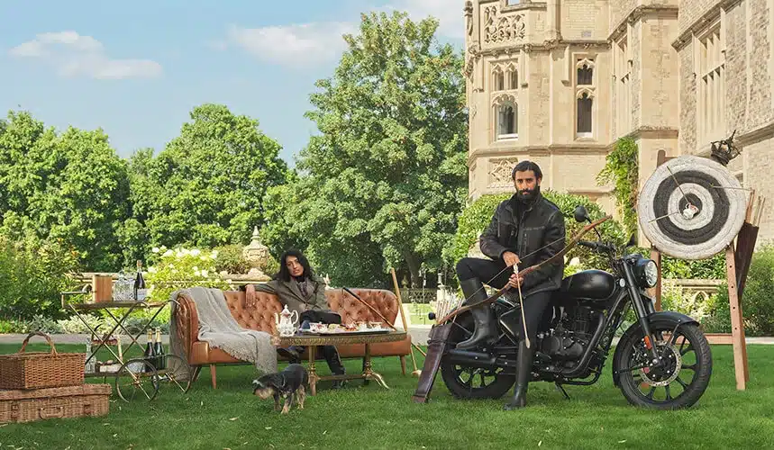 Royal Enfield Classic 350 Stealth Black vor großem Herrenhaus, daneben ein Paar auf Ledercouch und Bogenschieß-Accessoires