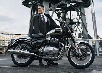 Royal Enfield Classic 650 Motorrad mit Chrom-Details und Peashooter-Auspuffen vor industriellem Hintergrund, Mann steht daneben.