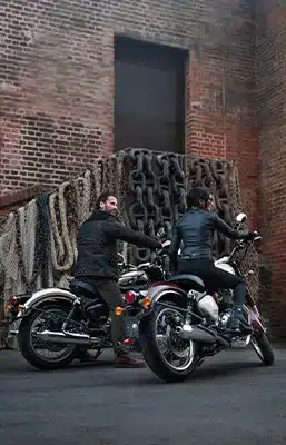 Zwei Personen sitzen auf Royal Enfield Classic 650 Motorrädern vor einer Backsteinwand mit verstauten Ketten.