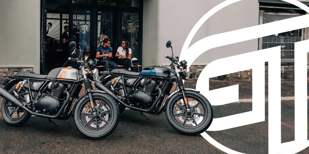 Zwei Royal Enfield Continental GT 650 Motorräder – eines in Silber mit orangen Akzenten, eines in Schwarz mit blauen Akzenten – parken vor einem modernen Café. Im Hintergrund sitzen zwei Personen auf einer Bank.