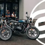 Zwei Royal Enfield Continental GT 650 Motorräder – eines in Silber mit orangen Akzenten, eines in Schwarz mit blauen Akzenten – parken vor einem modernen Café. Im Hintergrund sitzen zwei Personen auf einer Bank.