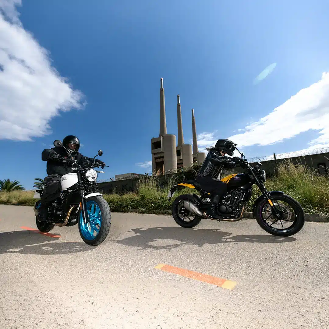 Zwei Royal Enfield Guerrilla 450 Motorräder auf städtischer Straße, im Hintergrund ein Industriegebäude mit Schornsteinen.