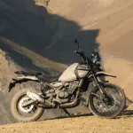 Seitenansicht einer Royal Enfield Himalayan 450 in beiger Lackierung, stehend auf einer staubigen Berghöhe vor felsiger Kulisse.