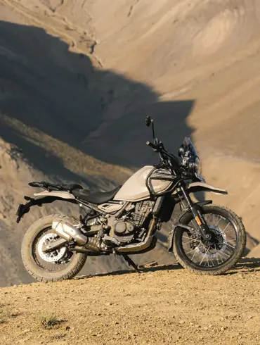 Seitenansicht einer Royal Enfield Himalayan 450 in beiger Lackierung, stehend auf einer staubigen Berghöhe vor felsiger Kulisse.