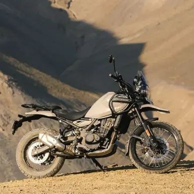 Seitenansicht einer Royal Enfield Himalayan 450 in beiger Lackierung, stehend auf einer staubigen Berghöhe vor felsiger Kulisse.