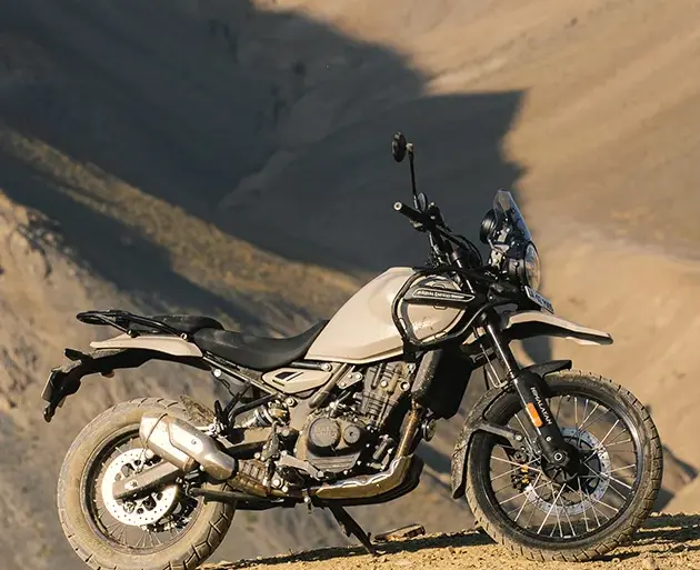 Seitenansicht einer Royal Enfield Himalayan 450 in beiger Lackierung, stehend auf einer staubigen Berghöhe vor felsiger Kulisse.