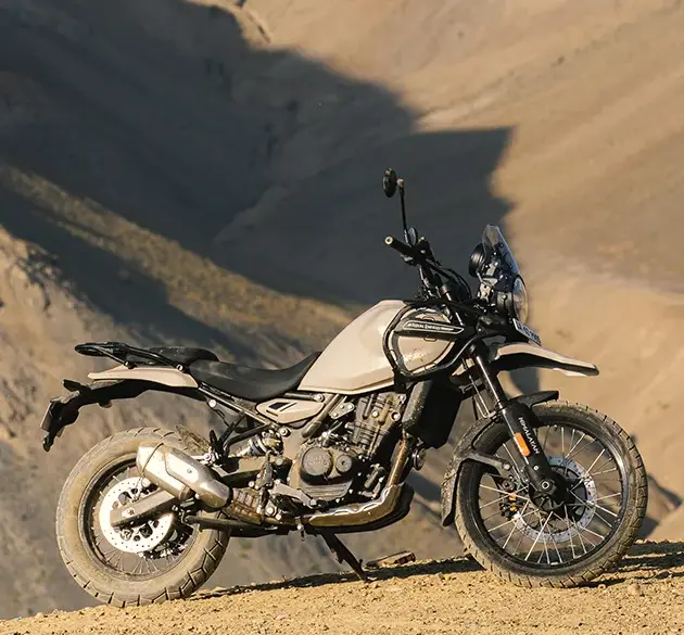 Seitenansicht einer Royal Enfield Himalayan 450 in beiger Lackierung, stehend auf einer staubigen Berghöhe vor felsiger Kulisse.