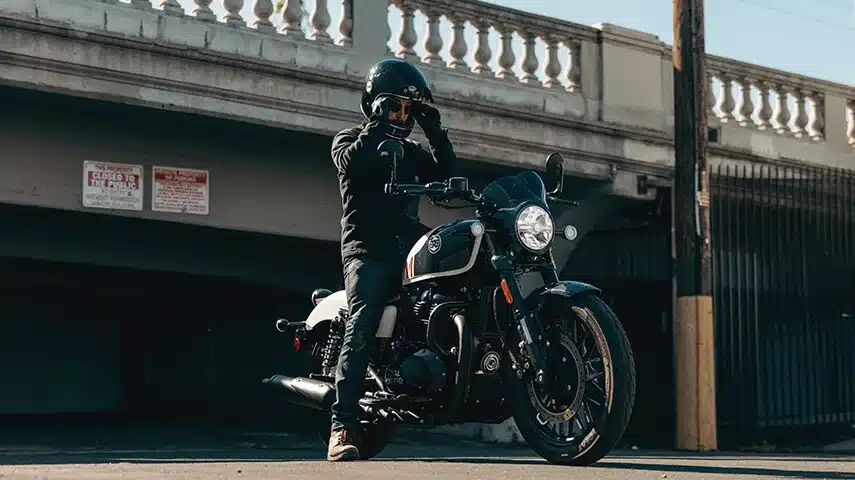 Fahrer mit Helm steht neben einer Royal Enfield Shotgun 650 vor einer Brücke.