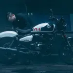 Ein Mann betrachtet sorgfältig eine weiß-schwarze Royal Enfield Shotgun 650 in einer stimmungsvoll beleuchteten Werkstatt.