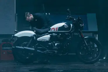 Ein Mann betrachtet sorgfältig eine weiß-schwarze Royal Enfield Shotgun 650 in einer stimmungsvoll beleuchteten Werkstatt.