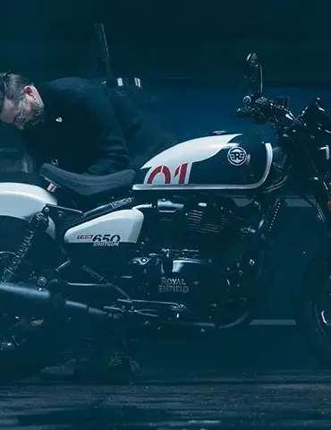 Ein Mann betrachtet sorgfältig eine weiß-schwarze Royal Enfield Shotgun 650 in einer stimmungsvoll beleuchteten Werkstatt.