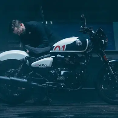 Ein Mann betrachtet sorgfältig eine weiß-schwarze Royal Enfield Shotgun 650 in einer stimmungsvoll beleuchteten Werkstatt.