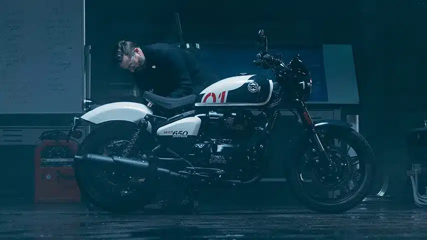 Ein Mann betrachtet sorgfältig eine weiß-schwarze Royal Enfield Shotgun 650 in einer stimmungsvoll beleuchteten Werkstatt.