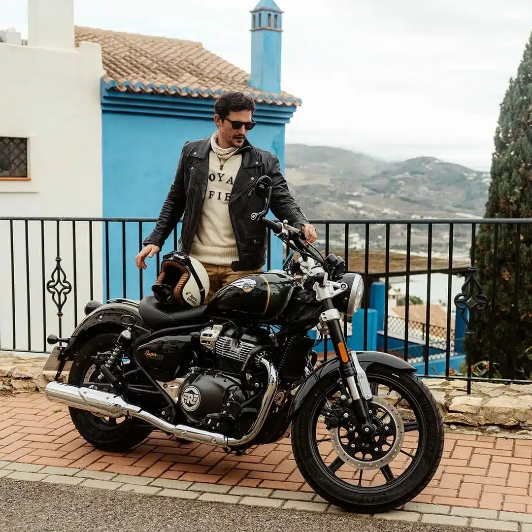 Royal Enfield Super Meteor 650 mit Fahrer vor Haus mit blauer Fassade und Hügellandschaft im Hintergrund.