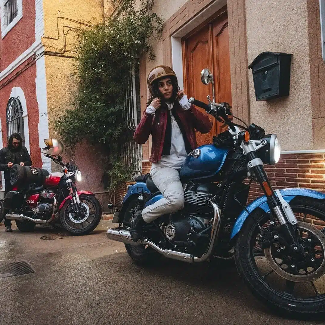 Royal Enfield Super Meteor 650 steht mit Fahrerin vor einem Haus, weitere Motorradfahrer im Hintergrund