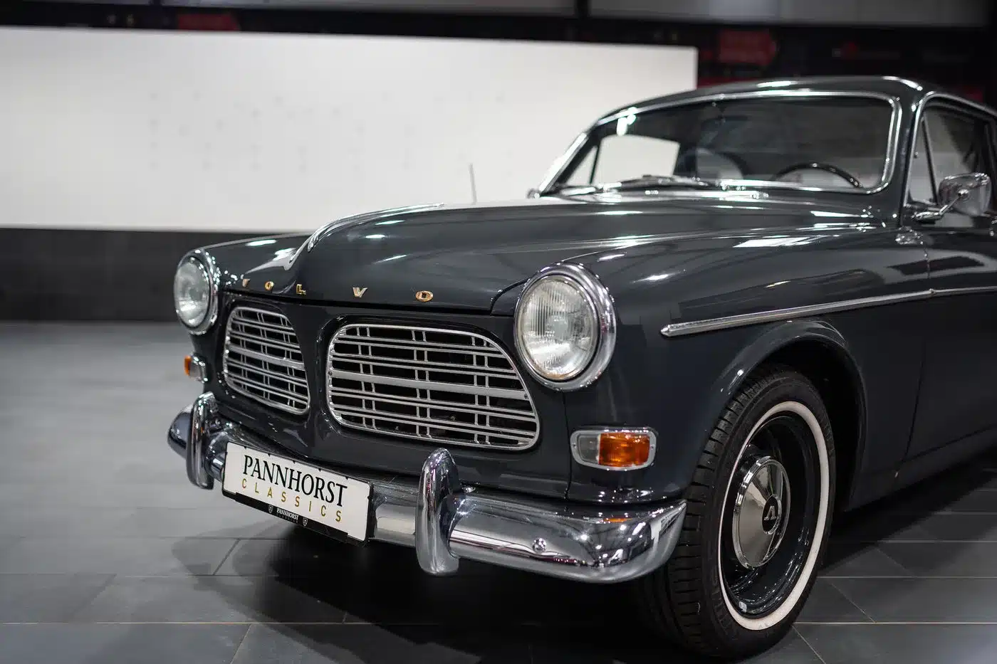 Frontalansicht eines dunkelgrauen Volvo Amazon Coupé, Baujahr 1964, mit verchromtem Kühlergrill und Stoßstange, aufgenommen in einer Ausstellungshalle.