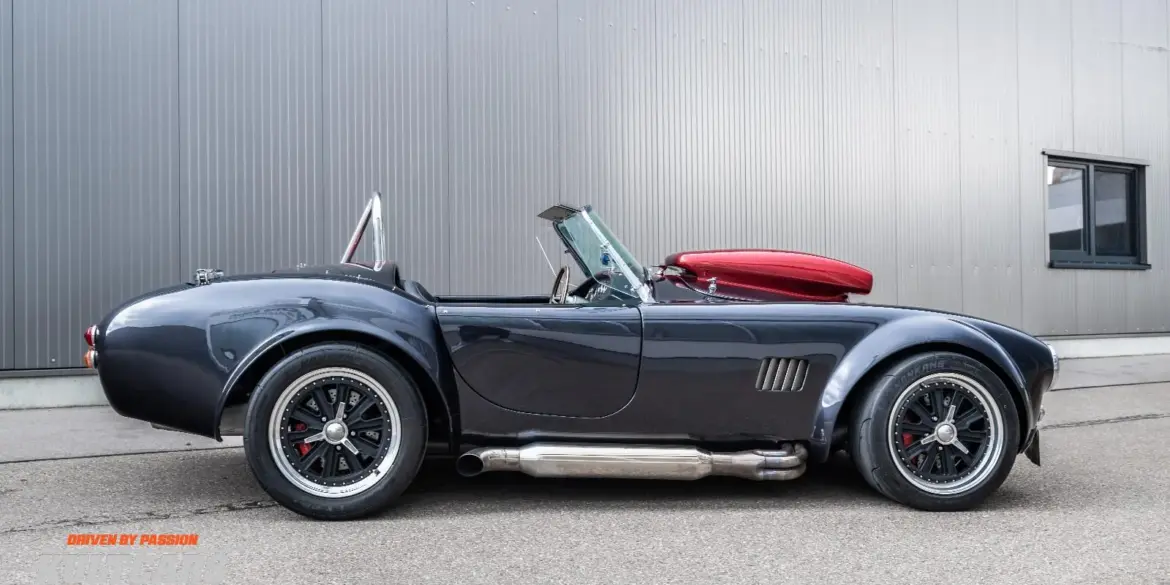 Graue Weineck Cobra 10.2L Cabrio in Seitenansicht vor industrieller Fassade, auffällige Sidepipes und rote Persenning.