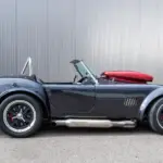 Graue Weineck Cobra 10.2L Cabrio in Seitenansicht vor industrieller Fassade, auffällige Sidepipes und rote Persenning.