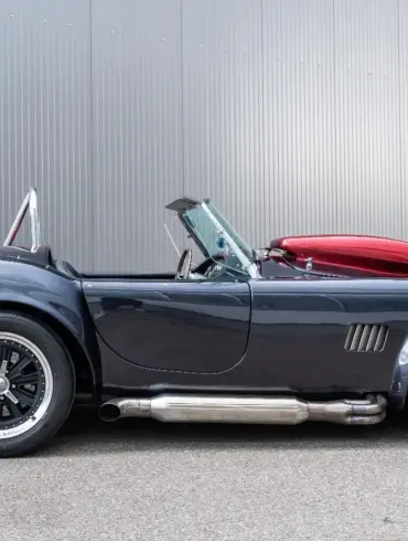 Graue Weineck Cobra 10.2L Cabrio in Seitenansicht vor industrieller Fassade, auffällige Sidepipes und rote Persenning.