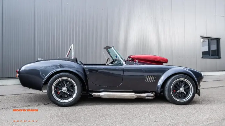 Graue Weineck Cobra 10.2L Cabrio in Seitenansicht vor industrieller Fassade, auffällige Sidepipes und rote Persenning.