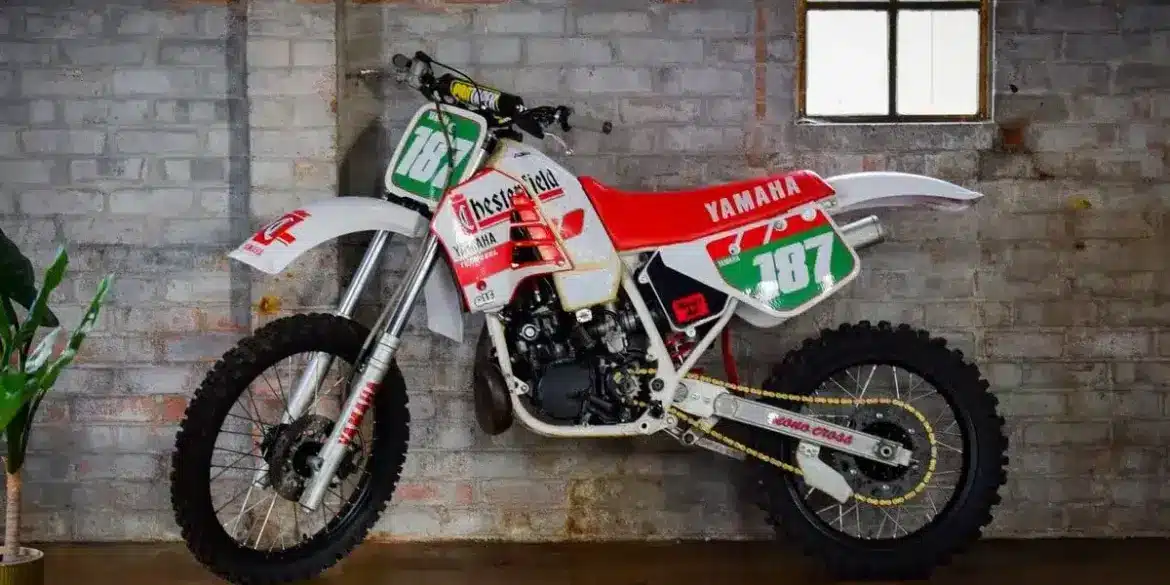 Seitliche Ansicht einer weißen Yamaha YZ250 (Baujahr 1987) mit Chesterfield-Grafiken, abgestellt vor einer Backsteinwand.