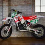 Seitliche Ansicht einer weißen Yamaha YZ250 (Baujahr 1987) mit Chesterfield-Grafiken, abgestellt vor einer Backsteinwand.