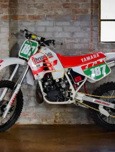 Seitliche Ansicht einer weißen Yamaha YZ250 (Baujahr 1987) mit Chesterfield-Grafiken, abgestellt vor einer Backsteinwand.