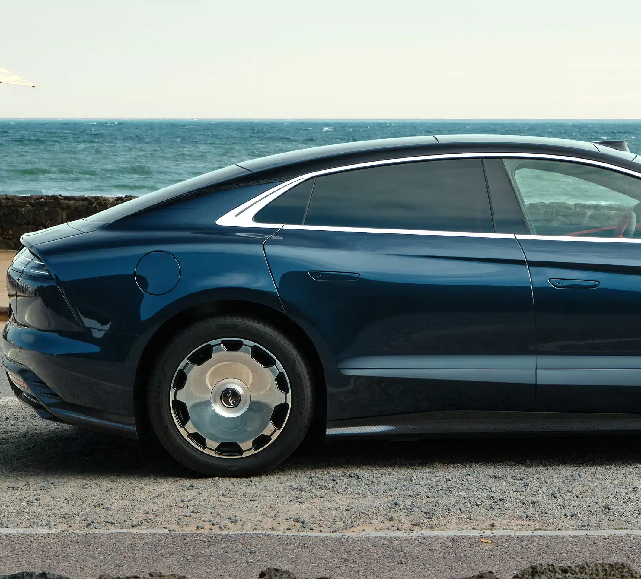 YangWang U7 Limousine in dunkelblau aus der Seitenperspektive, mit Fokus auf Heck und Hinterrad vor Meereskulisse.