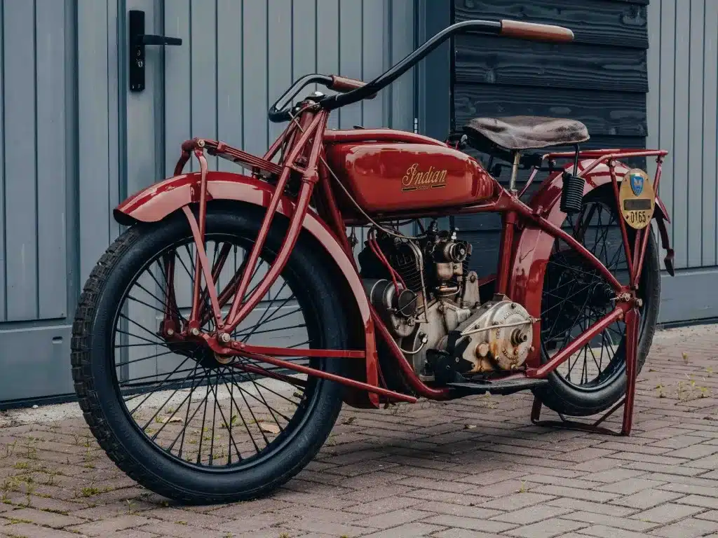 Rote 1925 Indian Scout, restauriertes Oldtimer-Motorrad mit V-Twin Motor, Girder-Vorderradgabel und Handschaltung, abgestellt vor grauen Garagentoren.