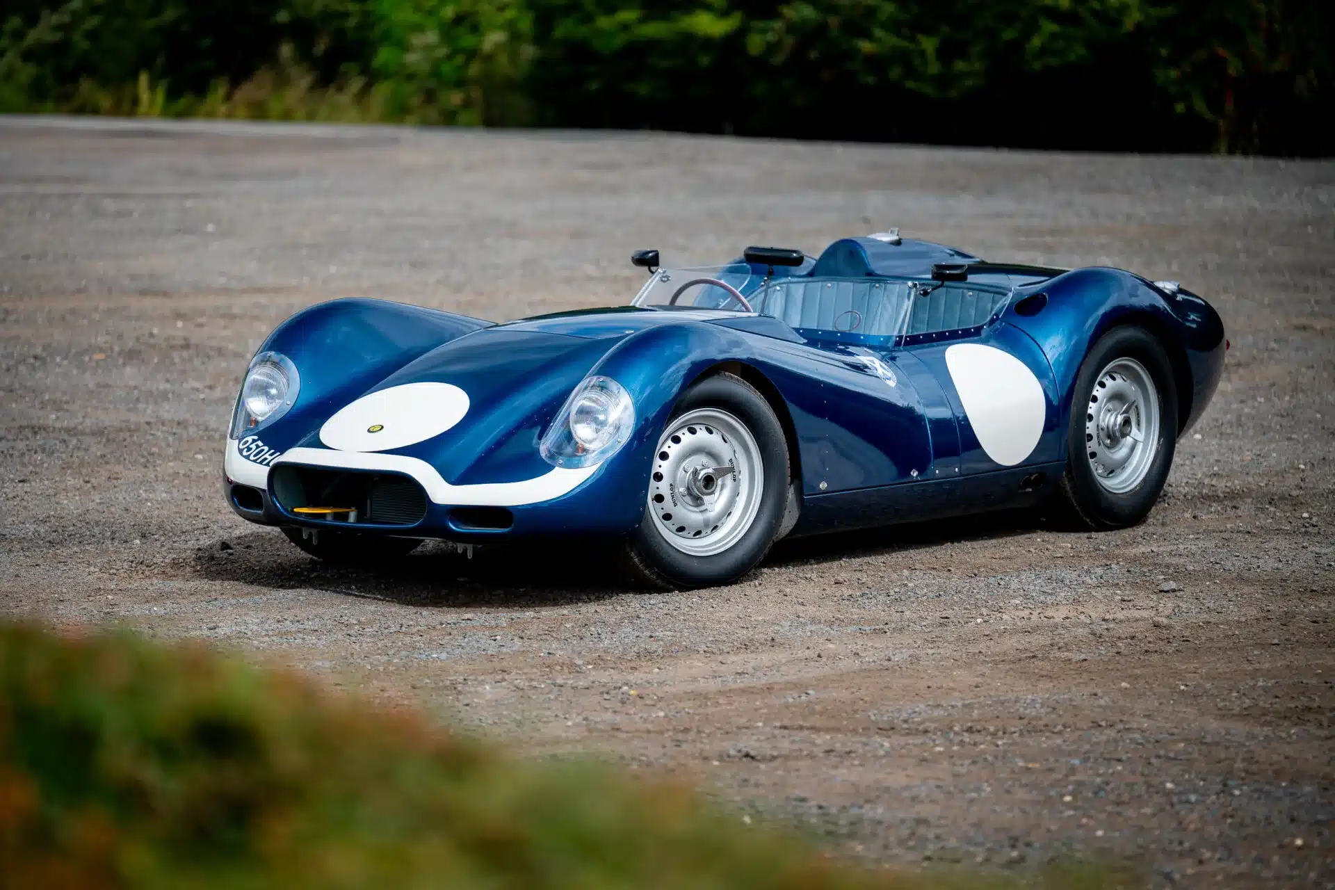 Blauer Lister-Jaguar Knobbly Continuation mit weißen Startnummernkreisen, fotografiert auf einem Schotterplatz, Front- und Seitenansicht.