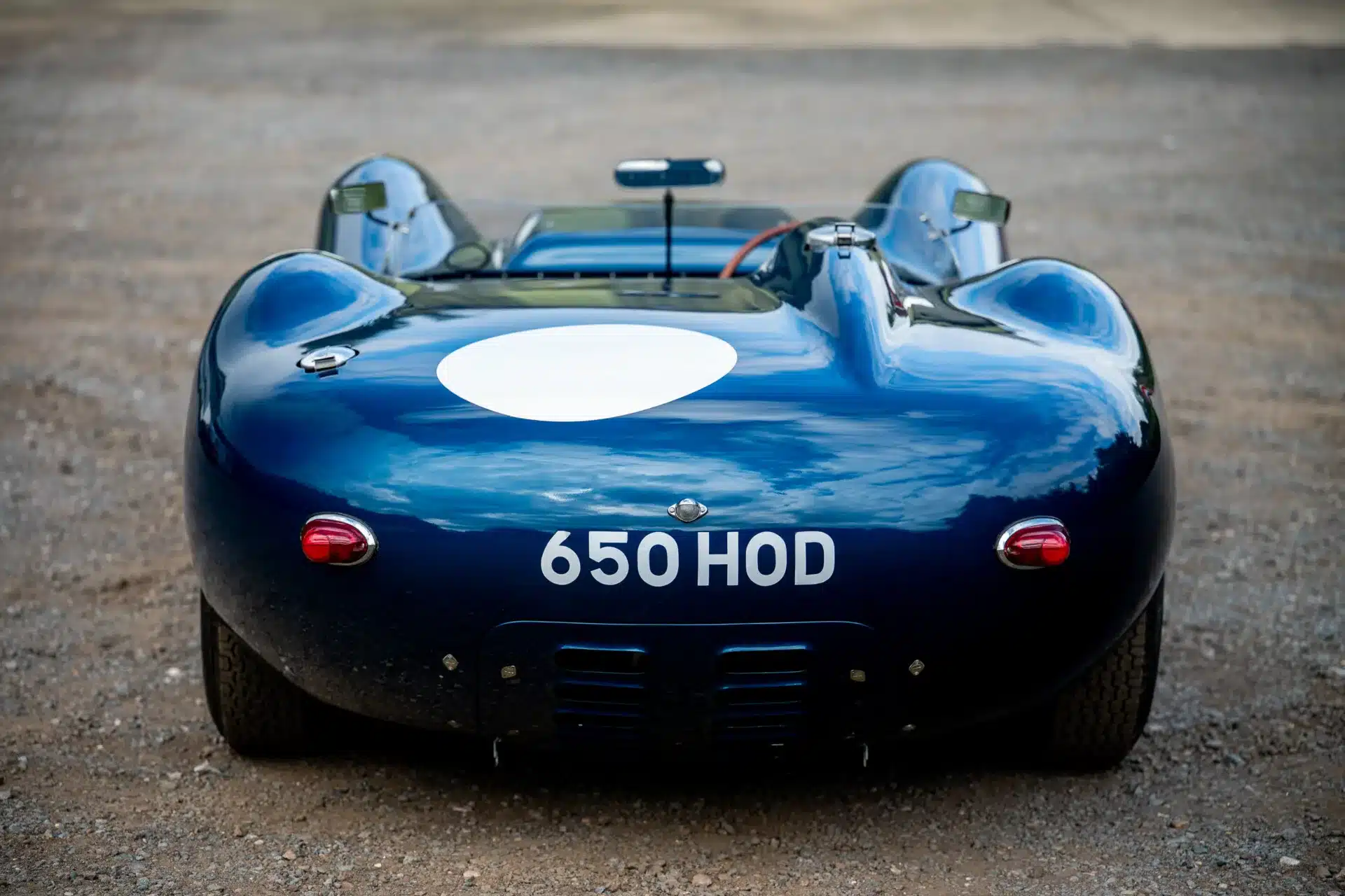 Lister-Jaguar Knobbly von hinten, lackiert in Ecurie-Ecosse-Blau mit weißem Kreis und der Aufschrift 650 HOD, auf einem Schotterplatz stehend.