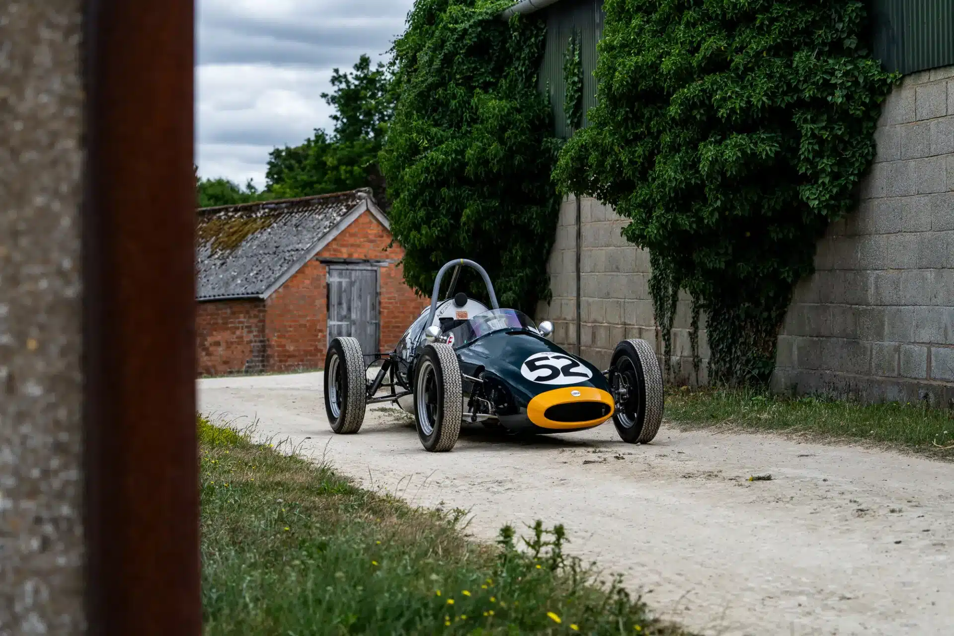 Cooper BMC T52 Rennwagen von 1960 mit Startnummer 52, dunkelgrüne Lackierung, fotografiert vor einer Backsteinscheune.