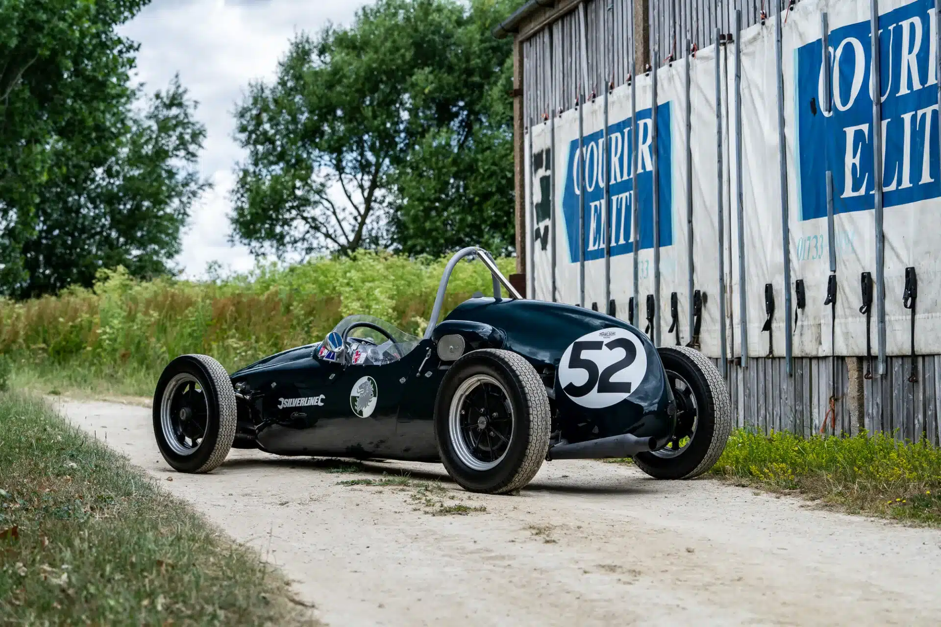 Cooper BMC T52 Rennwagen von 1960 mit Startnummer 52 in Dunkelgrün, geparkt auf einem Landweg vor einer Halle.