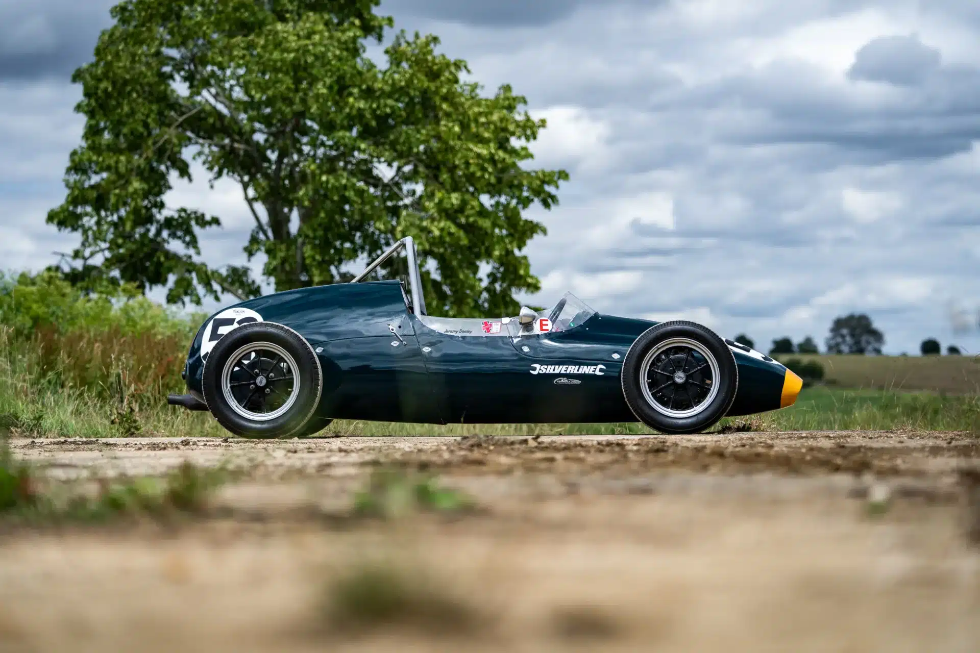 Seitenansicht eines dunkelgrünen 1960 Cooper BMC T52 Rennwagens auf einer Landstraße, im Hintergrund Bäume und bewölkter Himmel.