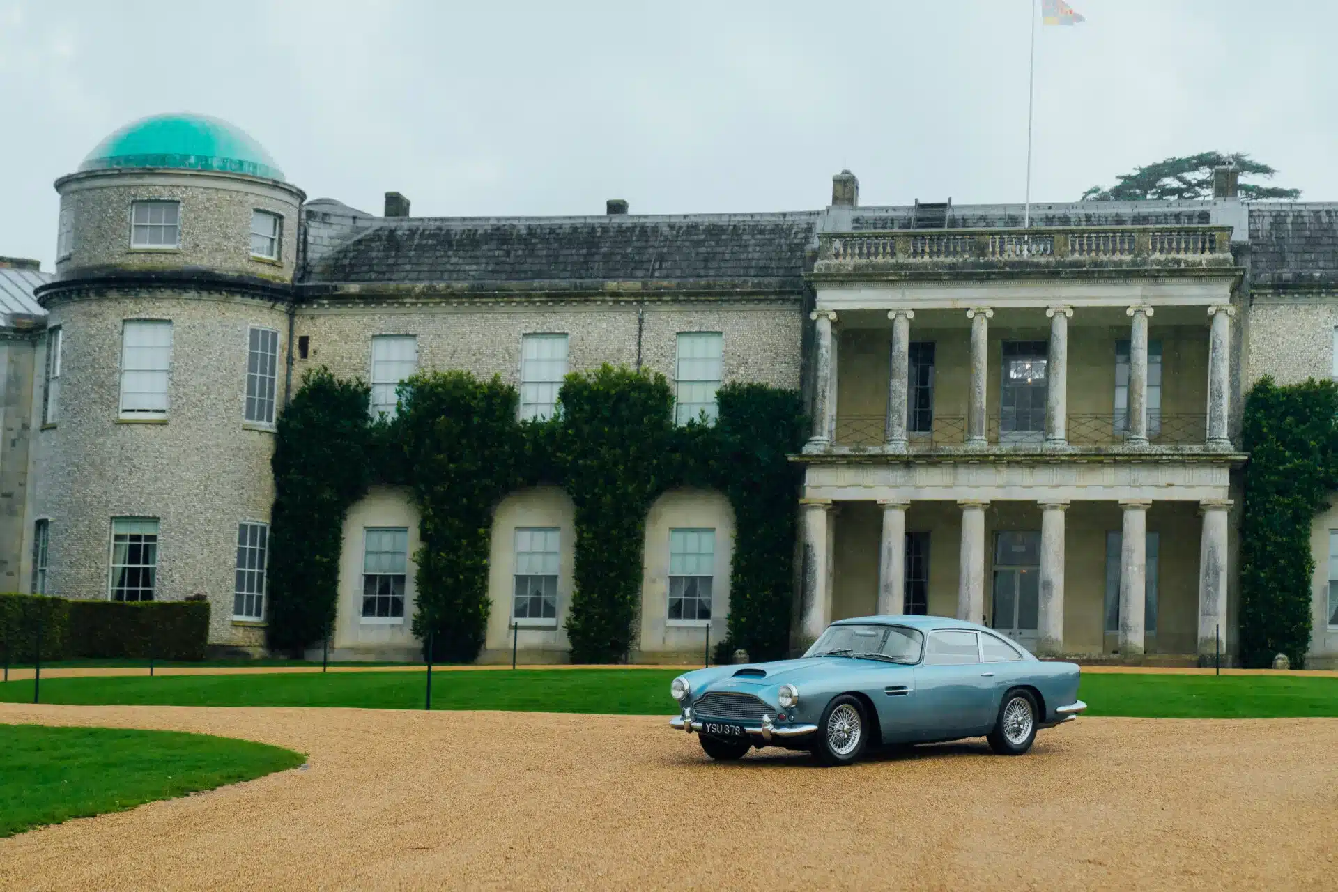 Aston Martin DB4 Series III Vantage von 1961 in karibikblauer Lackierung, geparkt auf Kies vor einem historischen Gebäude.